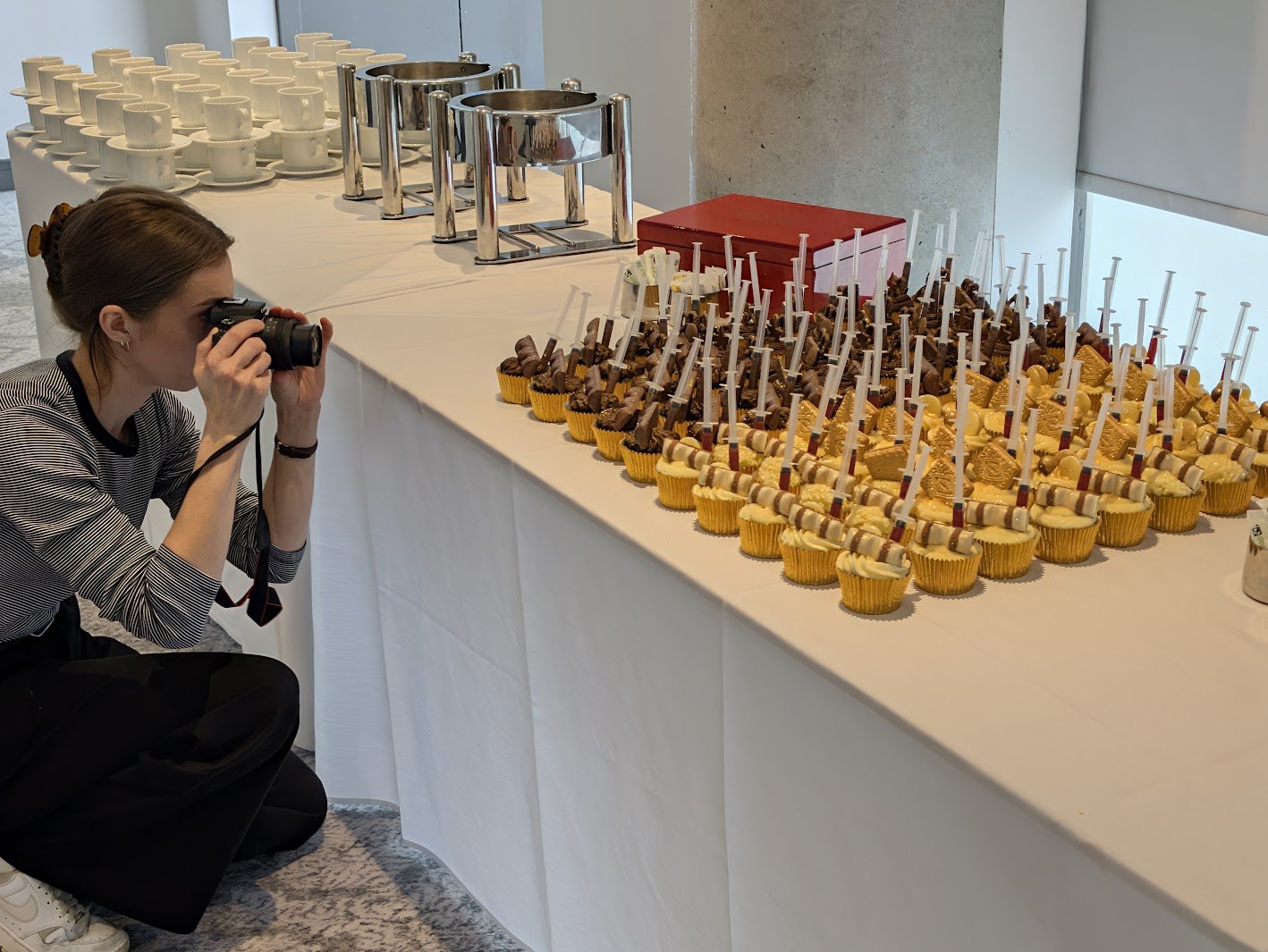 Abbie crouching beside a long white-clothed table, photographing rows of cupcakes topped with chocolate bars and toy syringes