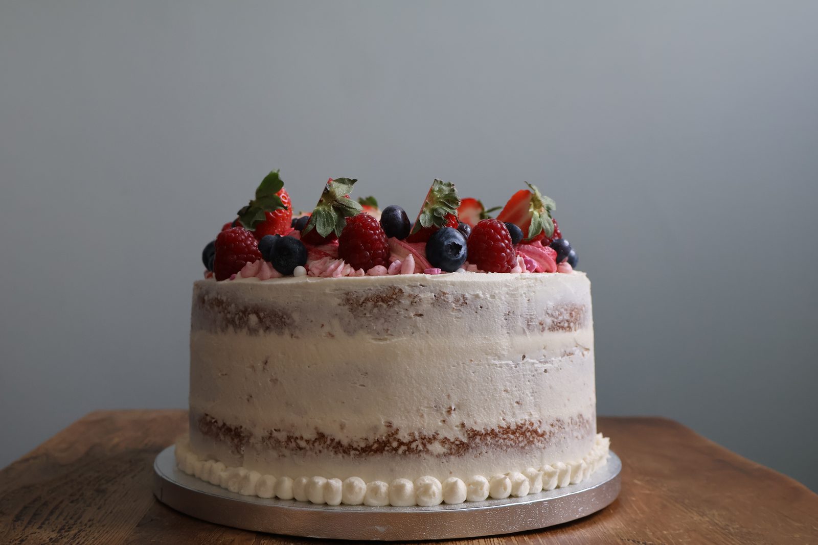 Semi-naked sponge cake with vanilla buttercream, topped with strawberries, raspberries and blueberries, on a wooden table